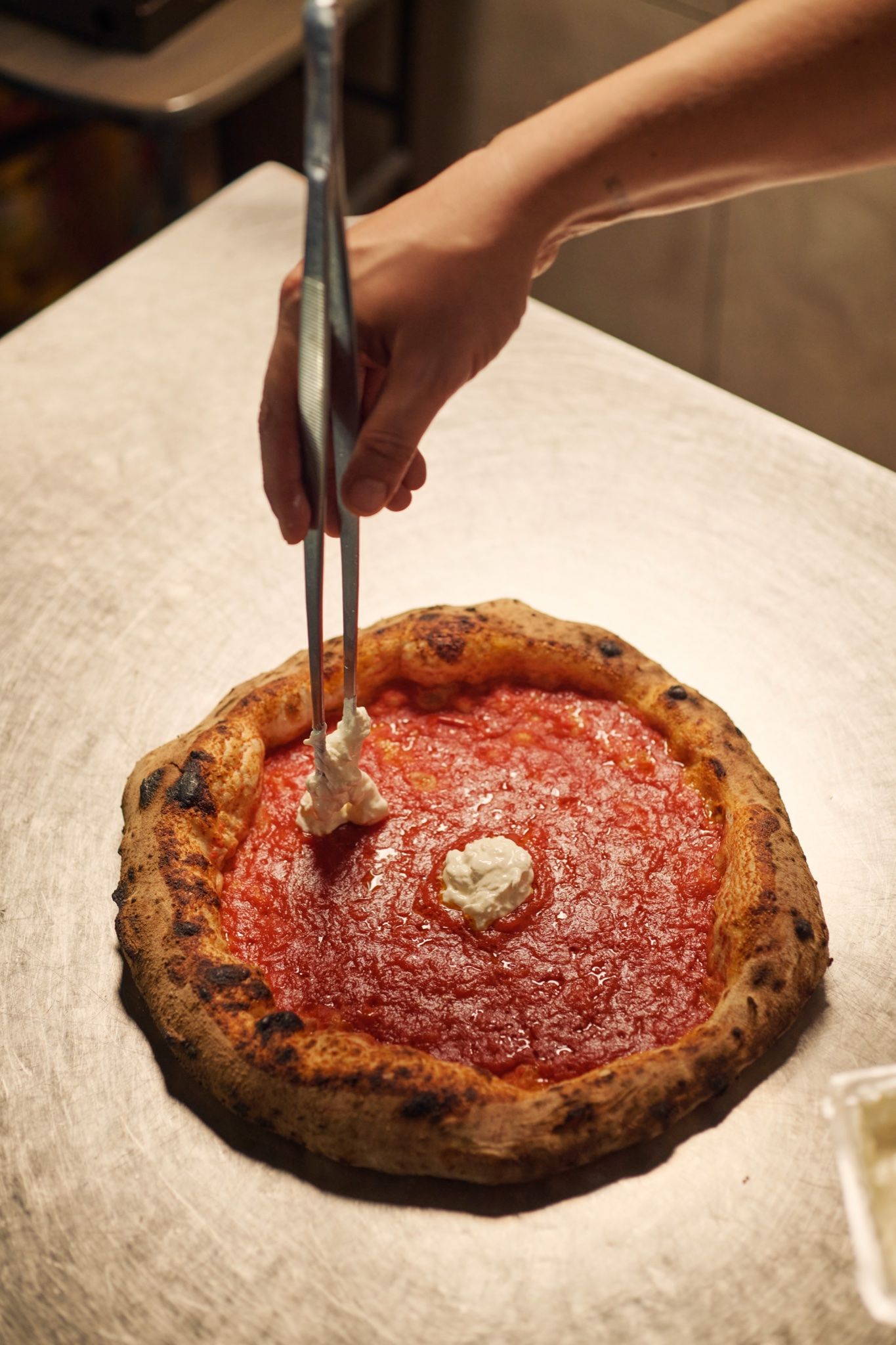 Una pinza posa la stracciatella sul pomodoro di una pizza appena sfornata